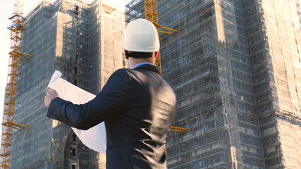 Portrait of a successful young handsome engineer, architect, builder, businessman, wearing a white helmet, in a suit, holding a project in his hand, a skyscraper background and a construction site.