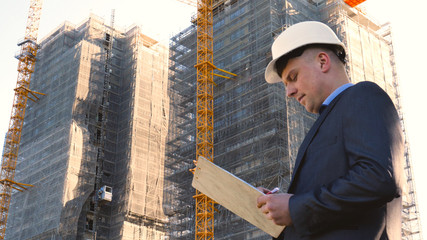 Portrait of a successful young handsome engineer, architect, builder, businessman, wearing a white helmet, in a suit, holding a notepad, skyscraper background and building.