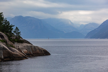 Rock goes into the calm water of misty Lisefjord with mountain slopes covered by blue haze on the background, Rogaland, Norway