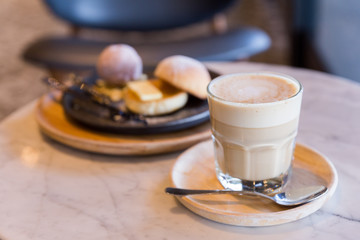 Hot latte in a glass cup with spoon and wooden plate with dessert in the background.
