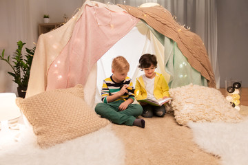 childhood, hygge and friendship concept - happy boys reading book with torch light in kids tent or...