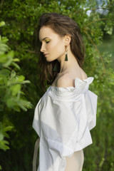 Beautiful stylish young girl wearing a trendy white shirt, beige trousers and a pareo poses outdoors against a background of green leaves. Nude makeup