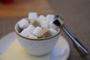 Close up white and brown sugar cubes in a bowl with a metallic pincers at the restaurant