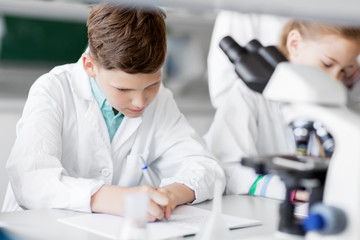 education, science and children concept - kids studying chemistry at school laboratory and writing to workbooks