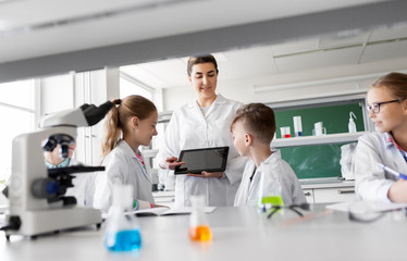 education, science and technology concept - chemistry teacher showing tablet pc computer to kids or students at school laboratory