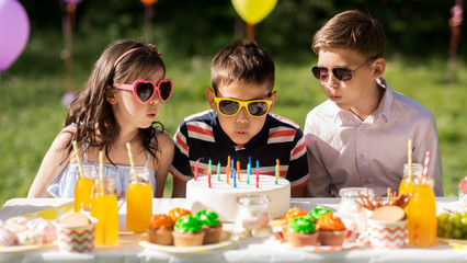 holidays, childhood and celebration concept - happy kids blowing out candles on birthday cake sitting at table at summer garden party