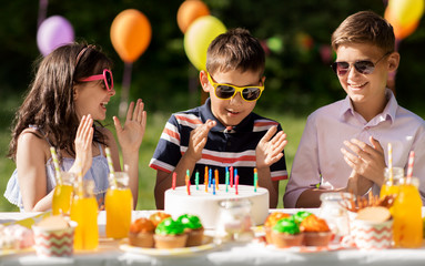 holidays, childhood and celebration concept - happy kids with candles on cake sitting at table at summer garden party and applauding to birthday boy
