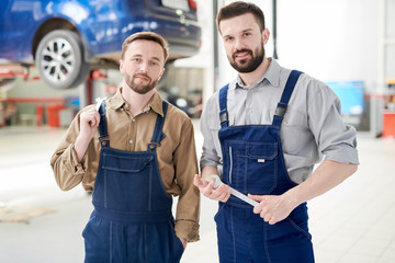Waist up portrait of two handsome bearded workers smiling at camera while posing with wrenches  in production workshop of modern car factory, copy space