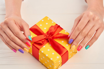 Colorful manicure and yellow gift box. Woman hands with colorful summer manicure holding gift box with red ribbon.