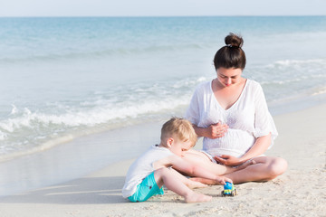 Portrait of happy pregnant mother relax near playing son at sea beach with white sand. Happy family at summer vacation concept