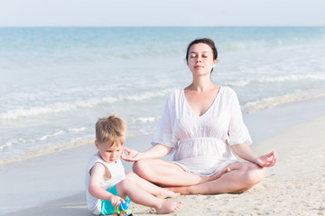 Young pregnant mother in white dress and her little toddler son playing and relaxing on the sea beach with sand at summer vacation time. Pregnancy, happy family, parenthood concept