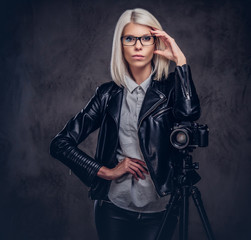 Smart blonde female photographer in trendy clothes posing while leaning on a professional camera with a tripod at a studio, looks at the camera.