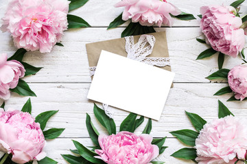 blank greeting card iand envelope in frame made of pink peony flowers over white wooden table with copy space