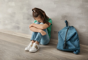 Upset little girl with backpack sitting on floor indoors. Bullying in school