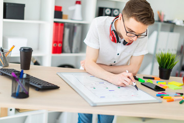 A young man in glasses stands near a computer desk. A young man draws a marker on a magnetic board. On the neck, the guy's headphones hang.