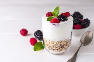 White yogurt with muesli and raspberries in glass bowl on white wooden background.