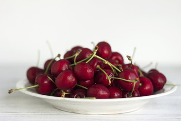 Ripe cherry berries with water droplets on a white plate on a white wooden background, clean, healthy and tasty fruit.
