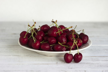 Ripe cherry berries with water droplets on a white plate on a white wooden background, clean, healthy and tasty fruit.