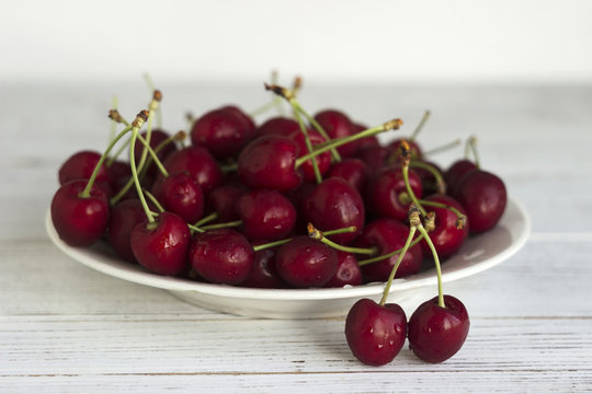 Ripe cherry berries with water droplets on a white plate on a white wooden background, clean, healthy and tasty fruit.