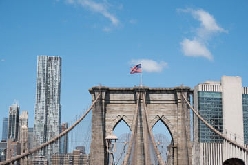 Naklejka premium View of lower Manhattan. Wall Street area, and the Brooklyn Bridge with American Flag on top from Brooklyn, New York. View of Manhattan's financial district and top of Brooklyn Bridge during the day.