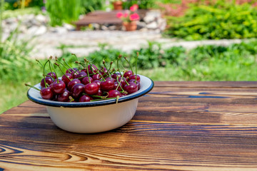 Many ripe red cherries in white metal bowl on the wooden table on open air. Close-up view