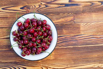 Many ripe red cherries in white metal bowl on the wooden table on open air. Top view. Copy space for text.