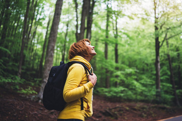 smiling tourist traveler with backpack into road at summer green forest, view back girl hiker in yellow hoody looking and enjoying the breath of fresh clean air in trip, relax holiday concept - obrazy, fototapety, plakaty