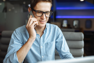 Portrait of a young man with a good mood, a businessman in a shirt and glasses, who works on a laptop and talking on the phone in a cafe, can be used for advertising, text insertion.