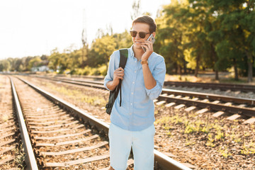 Beautiful stylish guy with glasses, posing on the train rails at sunset, and talking on the phone, hipster posing in stylish clothes, advertising, text insertion