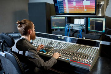 Stylish sound producer sitting at console and looking at monitors making up modern music. 