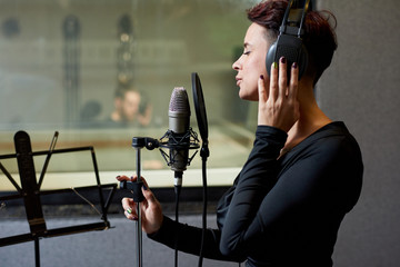 Side view of elegant woman in earphones singing with mic and recording song in studio. 