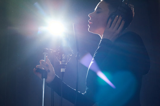From below side view of alluring girl wearing headphones while singing into microphone in bright light of recording studio. 