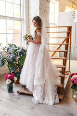 A bride with hairstyle and make up in gorgeous pink wedding dress and a vail with a bouquet of peonies. A portrait of beautiful girl with brunette hair and blue eyes in studio