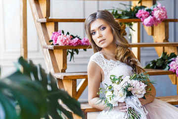 A bride with hairstyle and make up in gorgeous pink wedding dress and a vail with a bouquet of peonies. A portrait of beautiful girl with brunette hair and blue eyes in studio