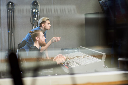 Side view through glass of creative operators sitting at music console in recording studio and talking to singer.