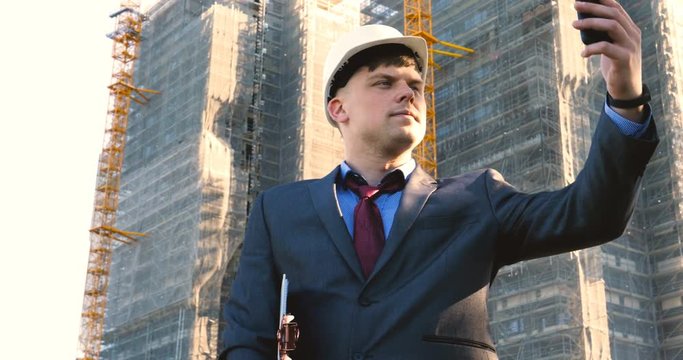 Portrait of a successful young handsome engineer, architect, builder, businessman, wearing a white helmet, in a suit, doing selfie, skyscraper background and building.