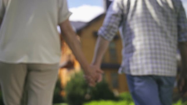Close-up of elderly couple's hands holding house door key and walking together on frontyard green lawn towards their new big dream house. Happy senor couple retired and purchased new home