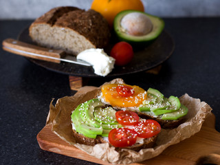 A delicious sandwich of whole-wheat bread and cream cheese, with avocado, cherry tomatoes, tomato and sesame, on a paper napkin on the background of products from which cooked. Healthy healthy food.