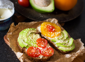 A delicious sandwich of whole-wheat bread and cream cheese, with avocado, cherry tomatoes, tomato and sesame, on a paper napkin on the background of products from which cooked. Healthy healthy food.