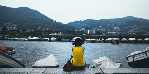 Young woman wearing yellow shirt and a black hat sitting at ease by the lake during sunset time.