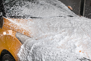 Dark yellow car being washed in carwash. Front completely covered in thick shampoo foam, with more...