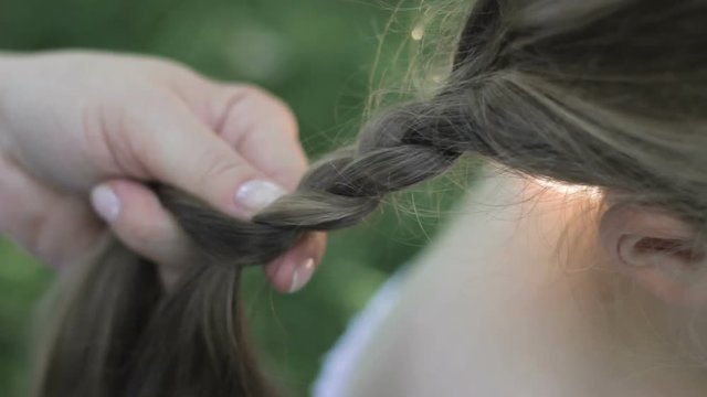 Oldish nanny's hands are braiding little child's hair, talking, resting outdoors