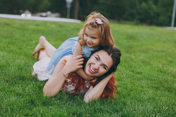 Fototapeta premium Laughing woman in light dress and little cute child baby girl hug, embrace and lie on grass rest and have fun. Mother, little kid daughter. Mother's Day, love family, parenthood, childhood concept.
