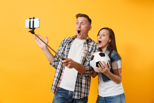 Young couple, woman man, football fans doing selfie on mobile phone with monopod selfish stick, cheer up support team, soccer ball isolated on yellow background. Sport family leisure lifestyle concept