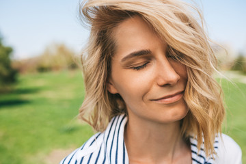 Close up outdoor portrait of pretty female with blond blowing hair and closed eyes enjoy weather. Close up shot of Caucasian beautiful young woman smiling with windy hair in park. People concept