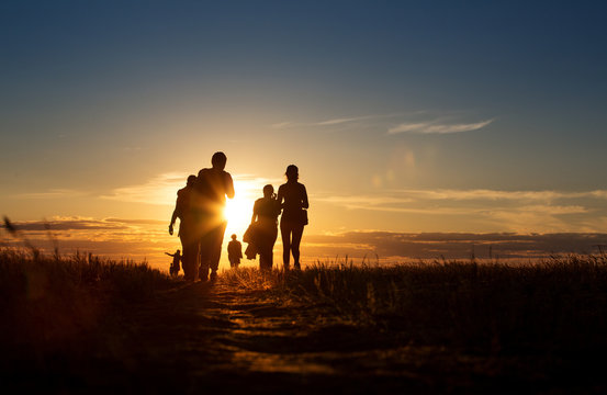 A group of people walking in a track. They go against the background of the orange sun, their contours and silhouettes are visible.
