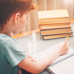 Red-haired boy writes in a notebook while doing school homework surrounded by books