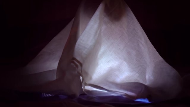 A child reads a book under blankets with a flashlight at night.