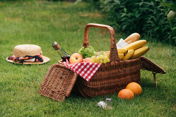 delicious fruits and bottle of champagne in wicker basket on green grass at picnic