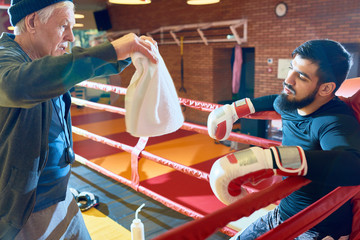 Side view of senior man training bearded young man sitting in ring corner and waving with towel for recreation